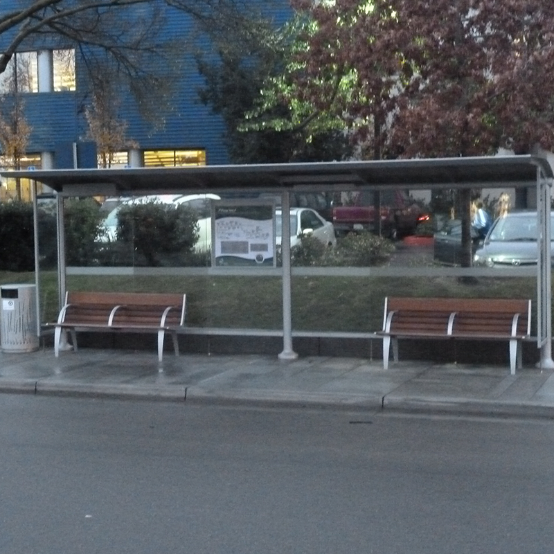 A front view of a bus shelter with two benches, one on either side, rider information displayed in the middle panel and a trash can nearby the left side
