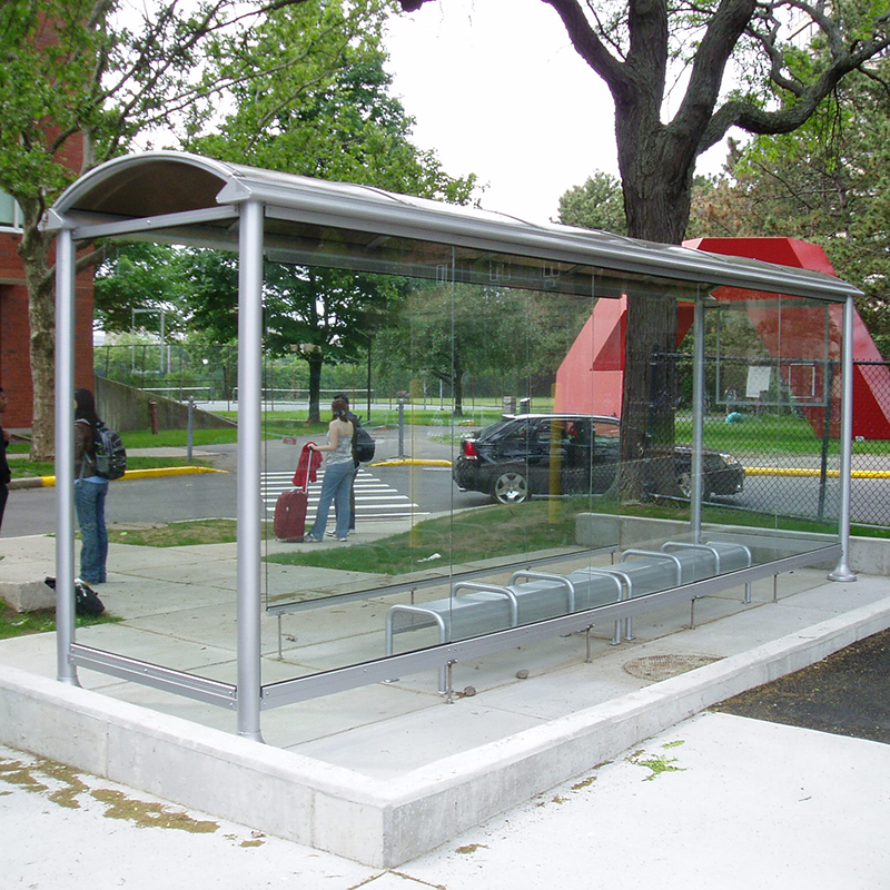 Riders are seen waiting for transportation outside a bus shelter with glass siding and ample bench seating inside the shelter.