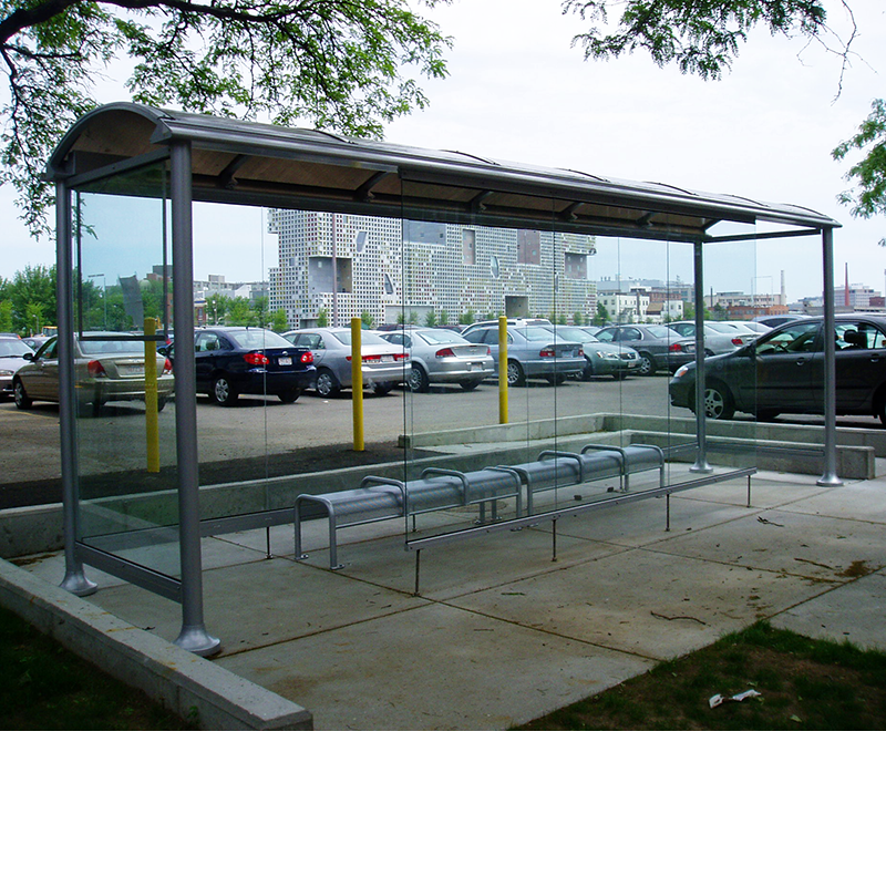 A bus shelter with glass paneling, bench seating for six seen in front of a parking lot.