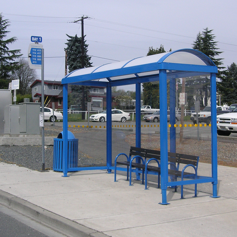 A bus shelter is seen with bench seating inside, a trash receptacle on the outside left and a bus stop sign with stop information next to the trash receptacle.