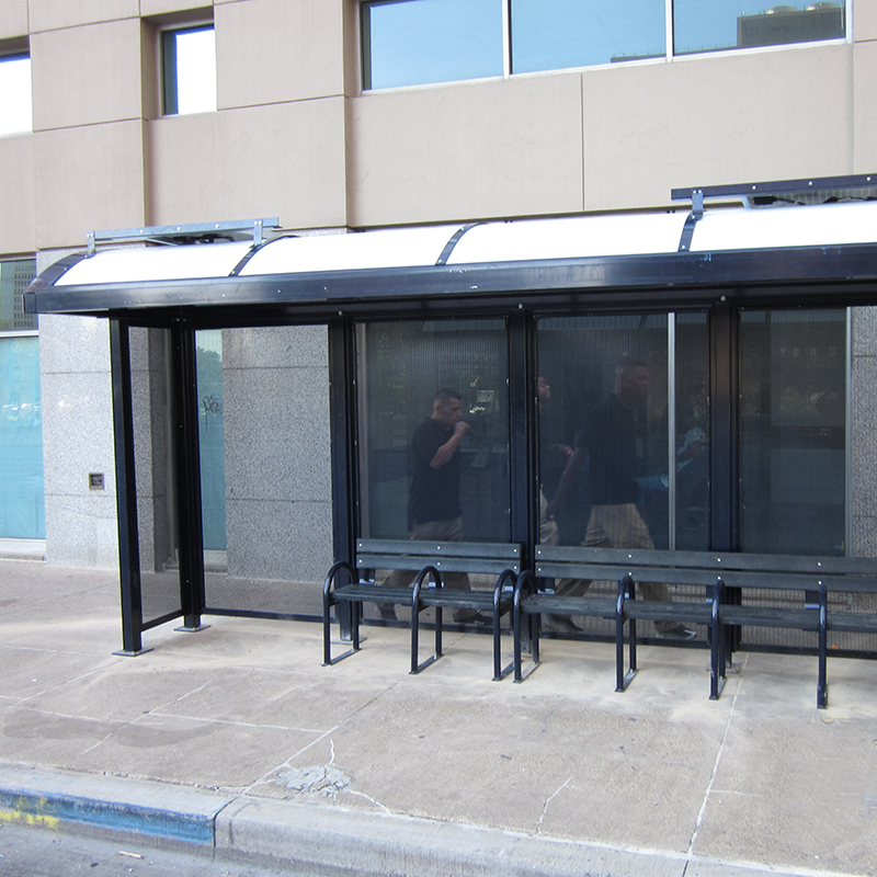 A bus shelter with two men walking behind it, solar panels are seen on the roof and bench seating for riders inside the shelter.