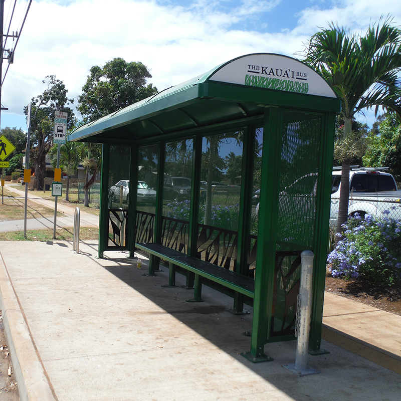 A bus shelter in Hawaii is seen with all green siding with a leaf grass design and a long bench for passengers as they wait.