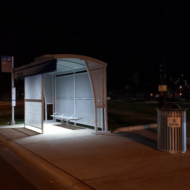 A left side view of a bus shelter at night with four sides, one bench and a trash can close by