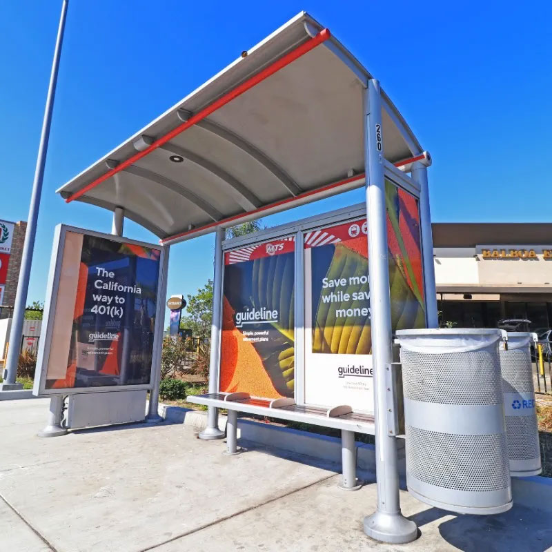 A bus shelter is seen from the front right side with 3 sides of the shelter having advertising displays, bench seating and two trash receptacles attached to the right side.