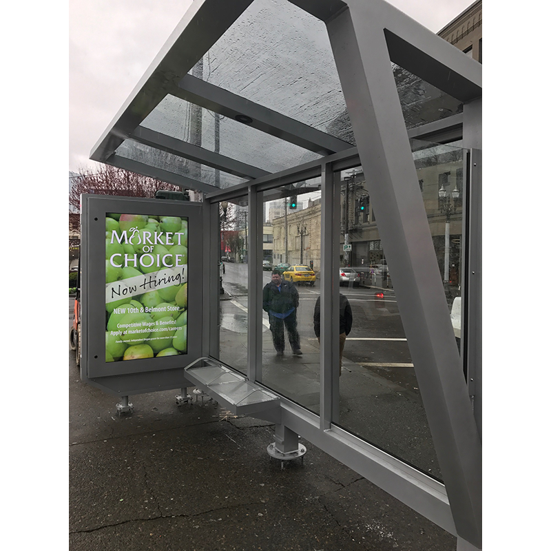 A bus shelter is seen from the right side with a left side panel advertisement for Market of Choice bench seating and a slanted roof with water running off of it from the rain.