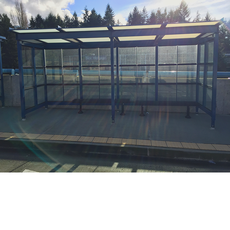 A bus shelter is seen from the front with bench seating inside and glass paneling as the sun shines through the back.