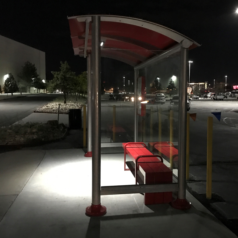 A bus shelter is seen from the right side at night with bench seating for three and overhead lighting inside the shelter.
