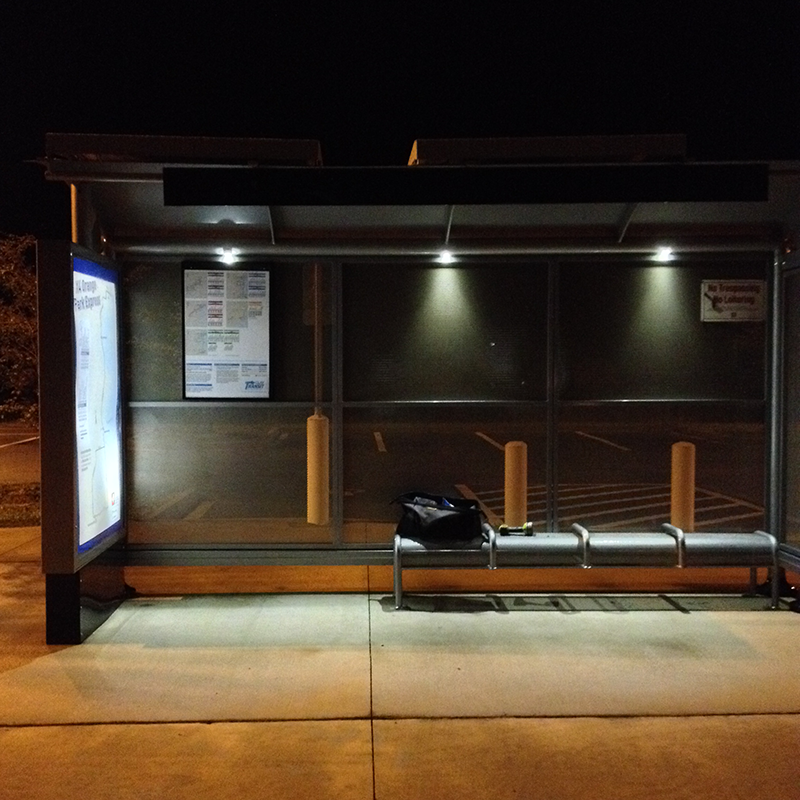 A bus shelter is seen at night from the front with the left side advertising display lit up, rider information and bench seating lit by the overhead lighting.