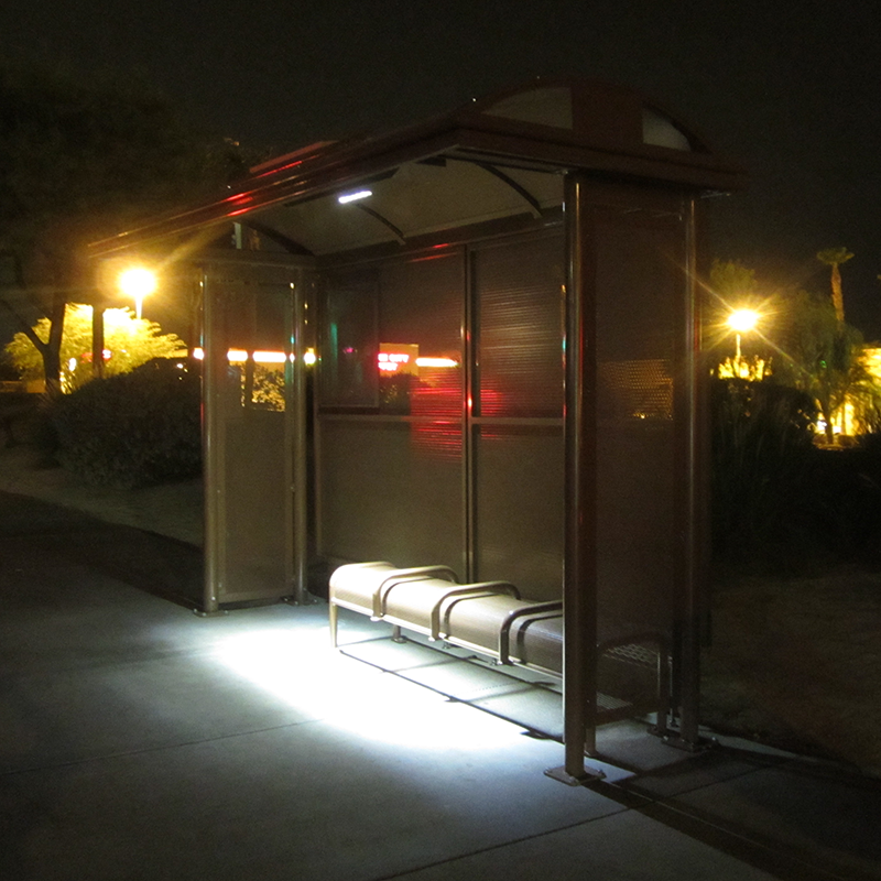 A bus shelter is seen at night with bench seating for five under a lit shelter.