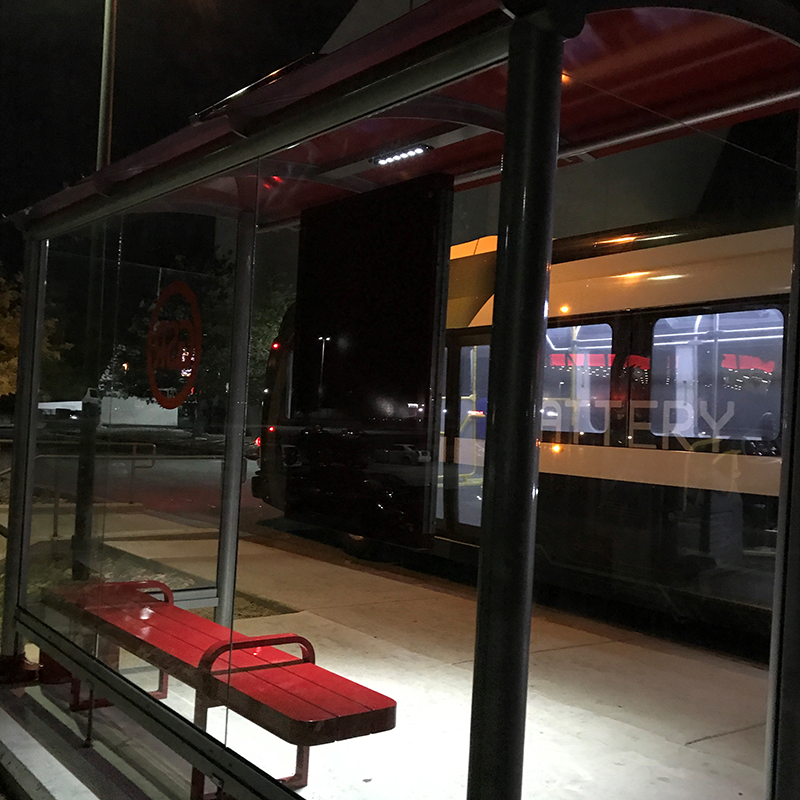 A bus shelter seen from the back right with glass siding that you can see through looking at the lit up bench seating at night and the bus that has pulled up to the stop.