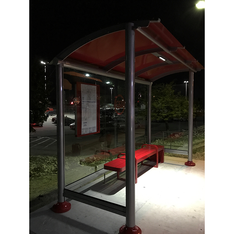 A bus shelter seen at night from the front left with bench seating inside and a rider information display on the back panel.