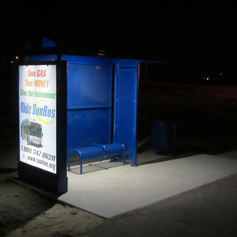 A bus shelter is seen from the front left with a double sided bus advertisement on the left side panel and bench seating inside the lit up shelter at night.