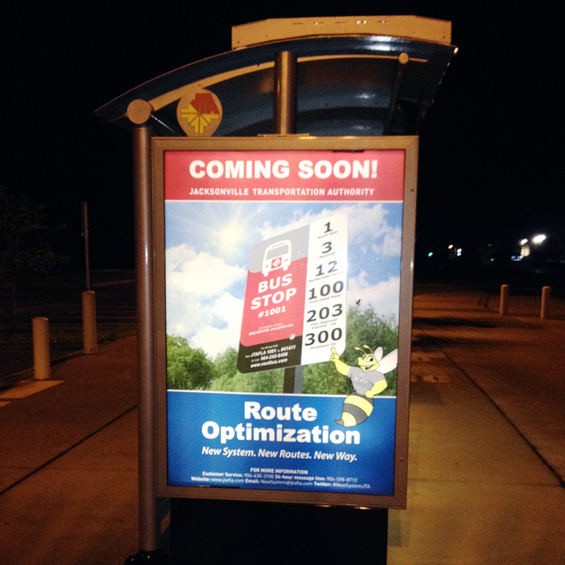 A bus shelter is seen from the left side with an advertising display lit up at night with a coming soon ad for the transportation company.