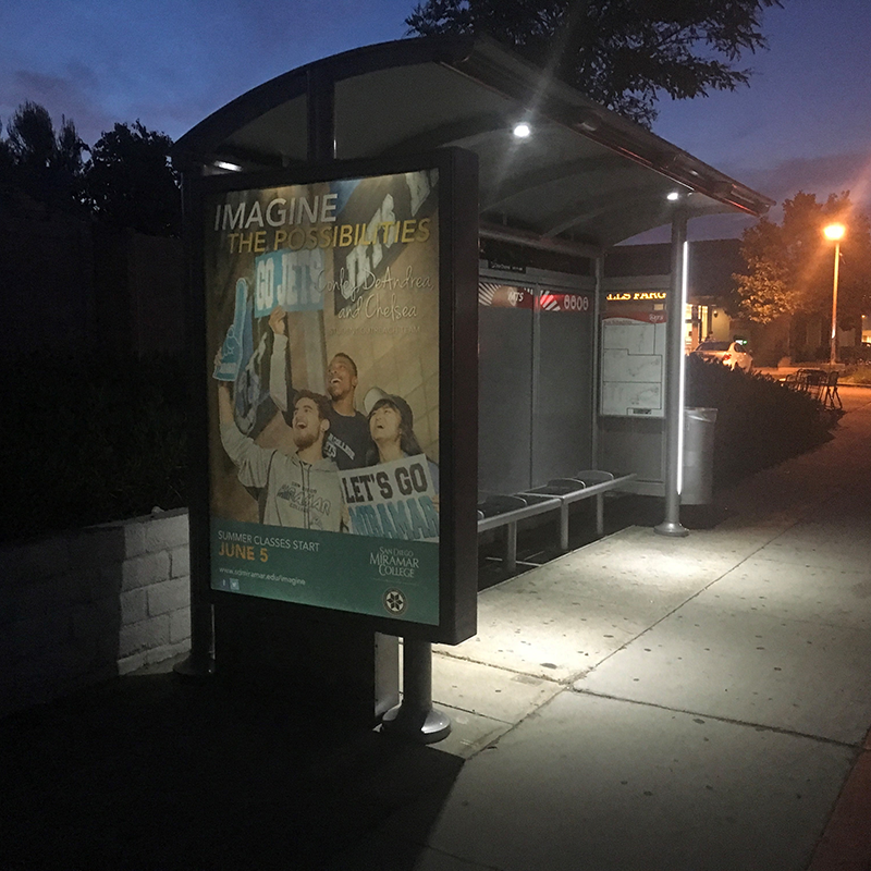A bus shelter is seen from the front left with an advertising display on the left side panel, bench seating inside, a rider information display on the right side panel and a trash receptacle attached to the outside right panel.