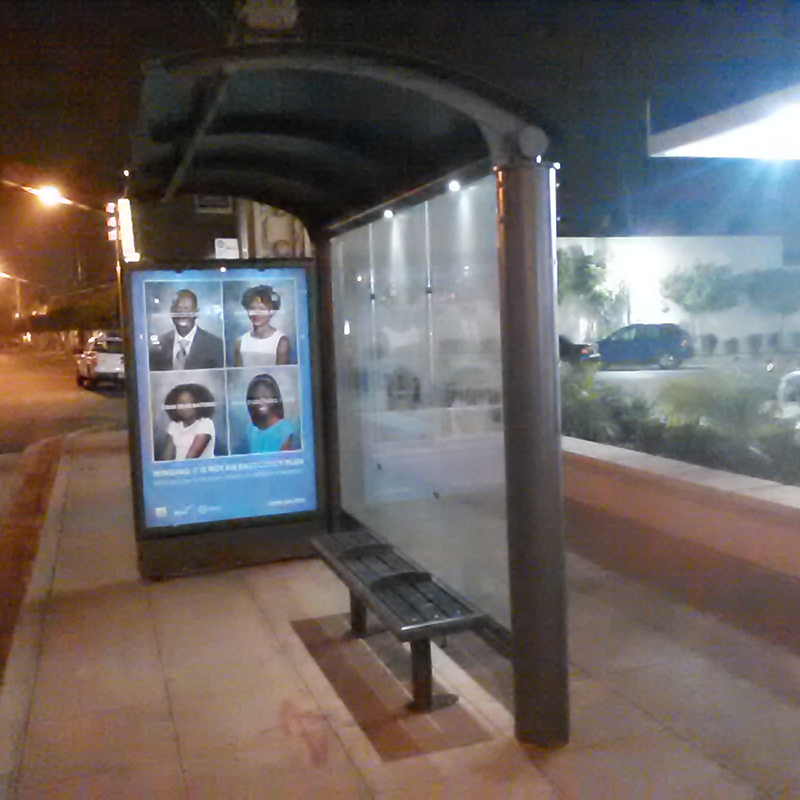 A bus shelter is seen from the right side with a left panel advertising display and bench seating inside.