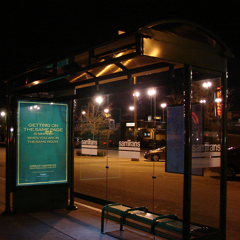 A bus shelter seen at night from the front right side with an advertising display on the left side, rider information on the back right panel and bench seating.