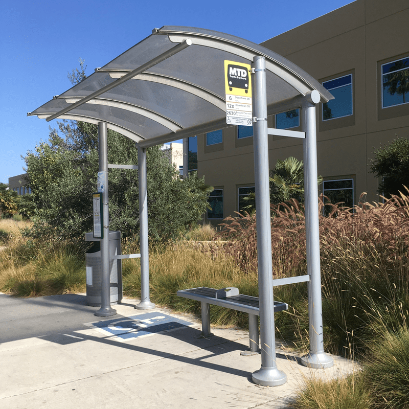 a bus shelter is seen from the front right with a left side trash receptacle outside the shelter and inside is a reserved spot for those in a wheel chair and bench seating.
