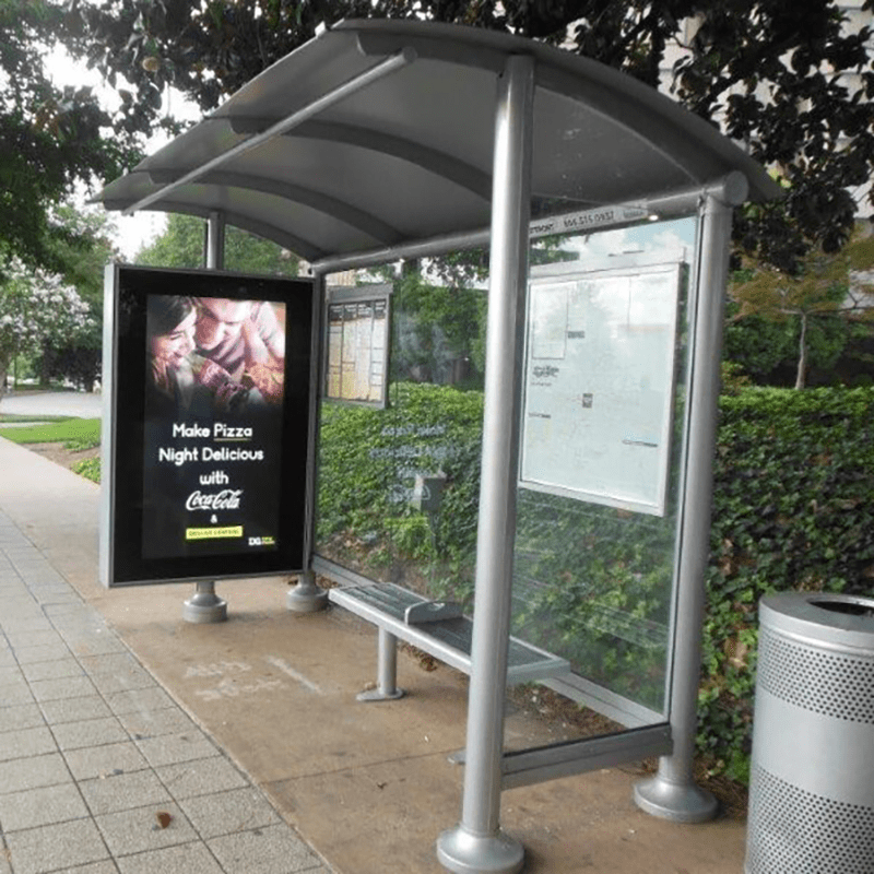 A bus shelter seen from the right side front with a trash receptacle on the right outside, bench seating, rider information display on the back panels and a left side panel for advertising.