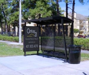 A bus shelter is seen from the front right with a left side panel advertising display, bench seating inside and a right side trash receptacle.