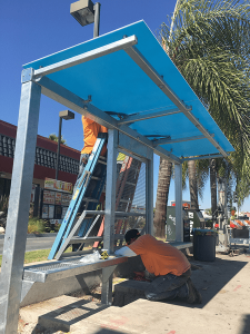 Workers are doing final touches on a bus shelter equipped with bench seating and paneling.