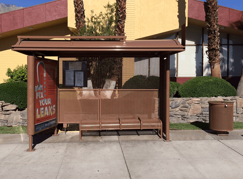 A front view of a brown bus shelter showcasing an advertising display on the right side, rider information postings along the back and bench seating