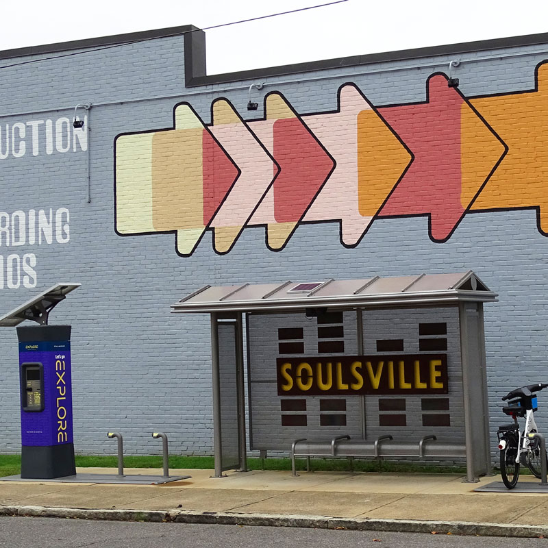 A bus shelter is seen in Soulsville with a custom back panel to the structure, bench seating and a ticket kiosk outside the shelter.