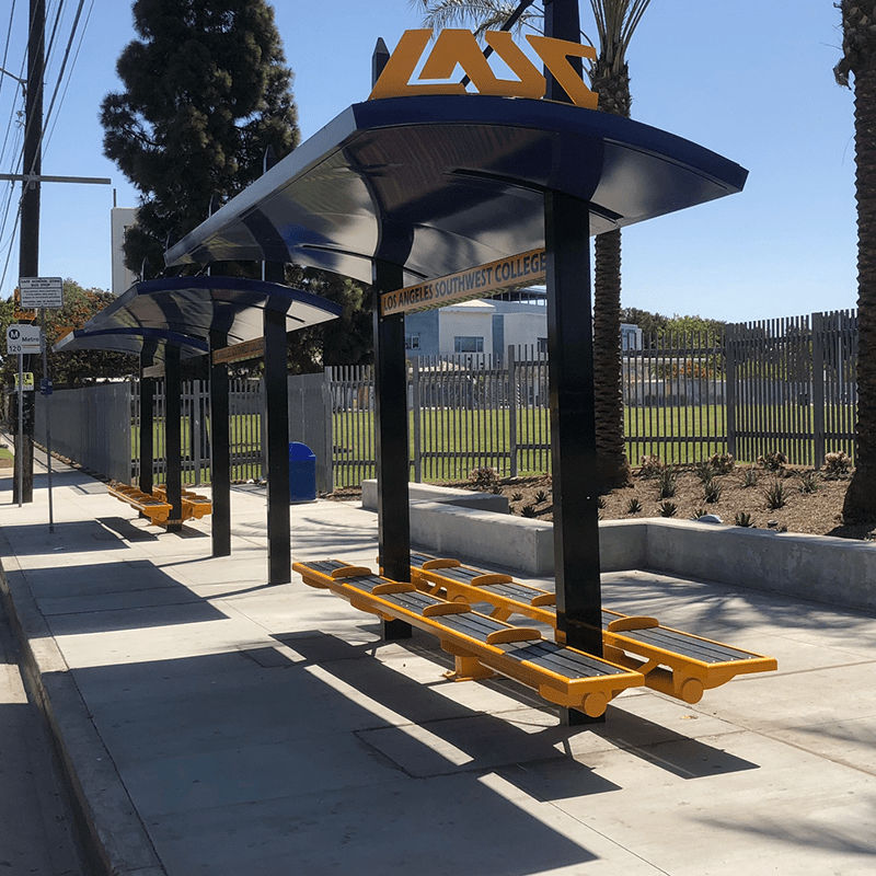 A signa bus shelter is seen from the right side with bench seating on either side of the structure and arched roofing.