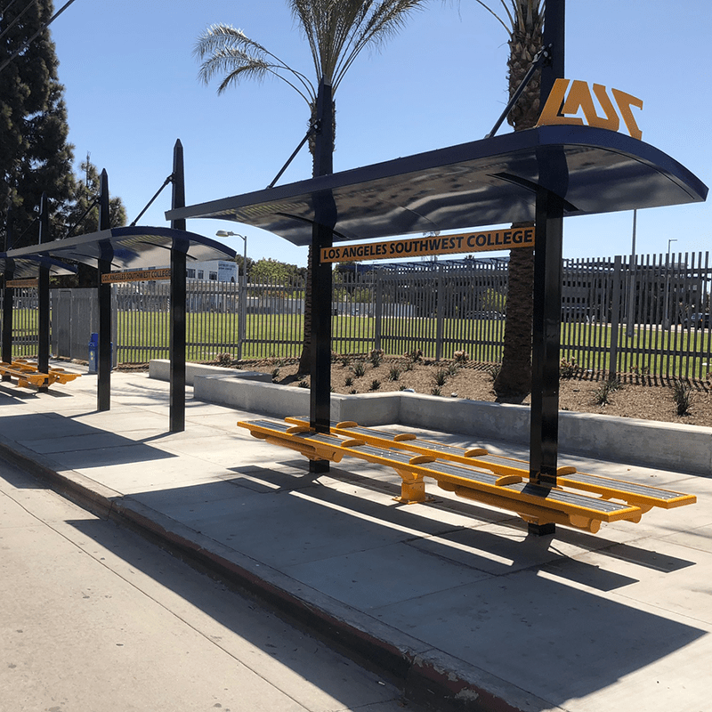 A bus shelter is seen from the front right with benches on either side of the structure posts outside of a Los Angeles Southwest College stop.