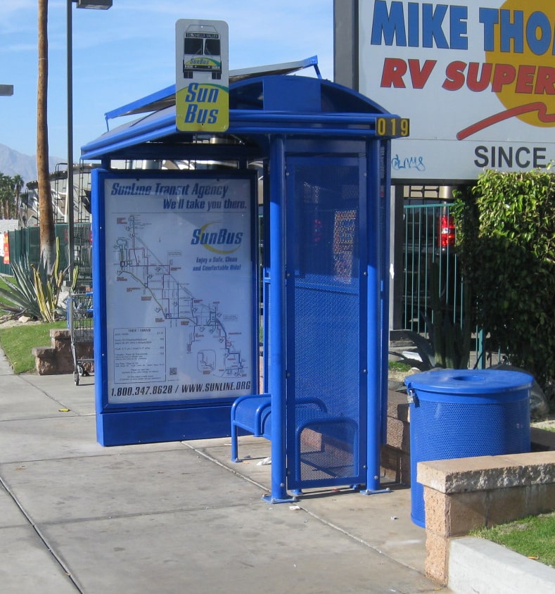 A bus shelter is seen from the front with a left panel advertising display, a trash receptacle on the right and bench seating inside the shelter.