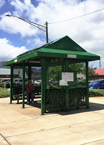 A bus shelter is seen from the right side with riders waiting inside on the benches provided facing inwards towards each other.