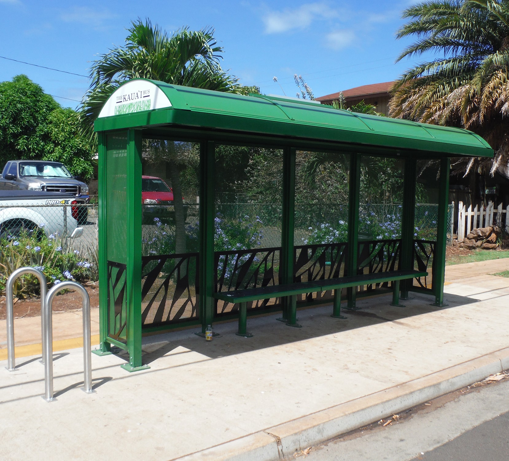 A bus shelter is seen with a leaf design on the bottom of the side and back panels with bench seating inside and bike stands on the outside left.