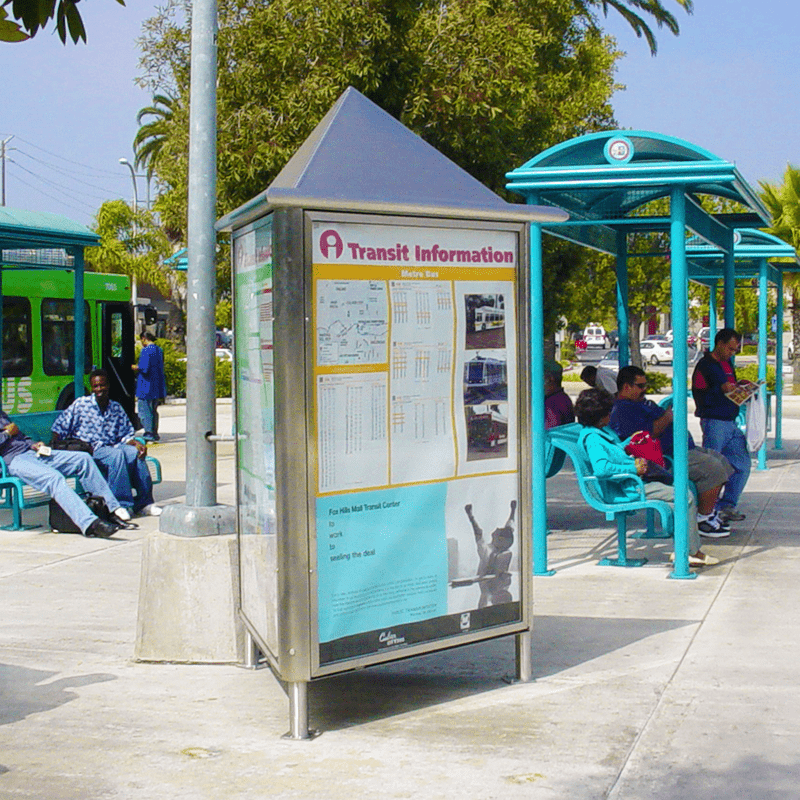 A rider information kiosk is seen at a transit center with full benches and a bus in the background.
