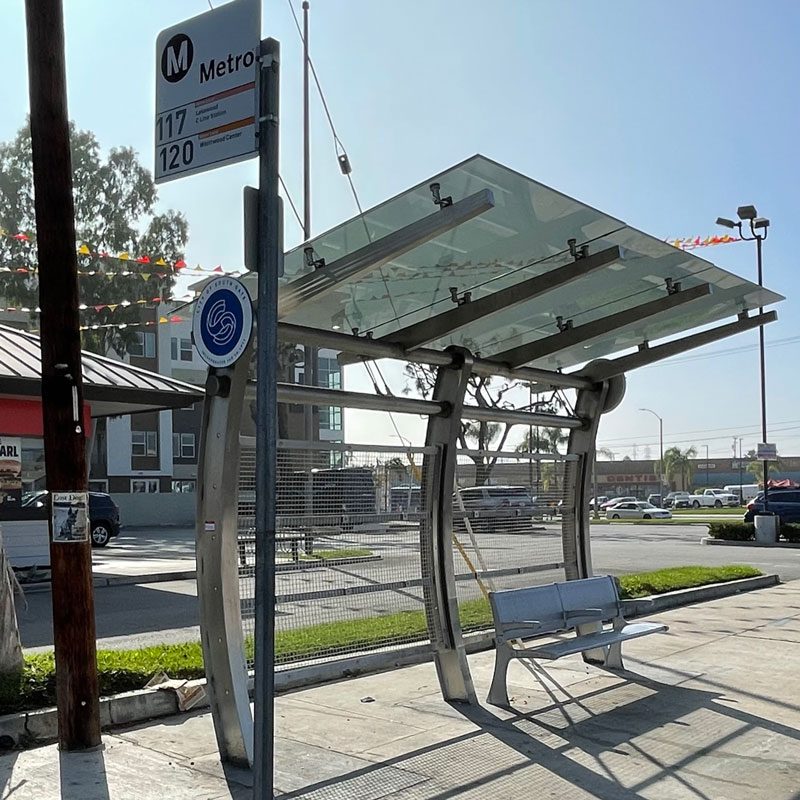A bus shelter in South Gate is seen from the front left side with an arched structure and bench seating for two under the shelter.