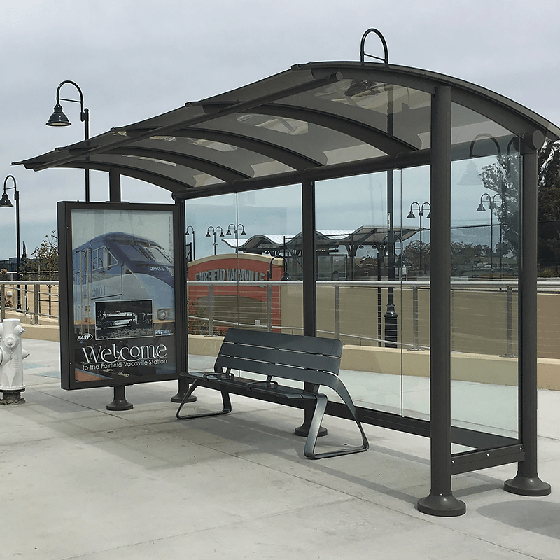 A siga sunset bus shelter is seen with glass siding, an advertising display on the left side panel and a bench for rider seating.