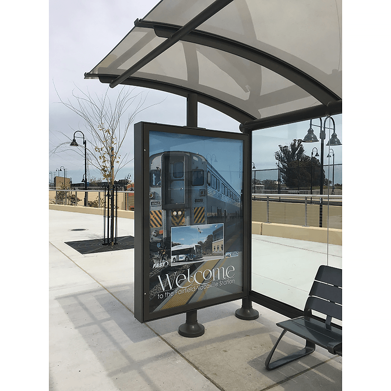 A siga sunset bus shelter's advertising display on the left side panel with a welcome to the Fairfield-Vacaville Station advertisement for riders.