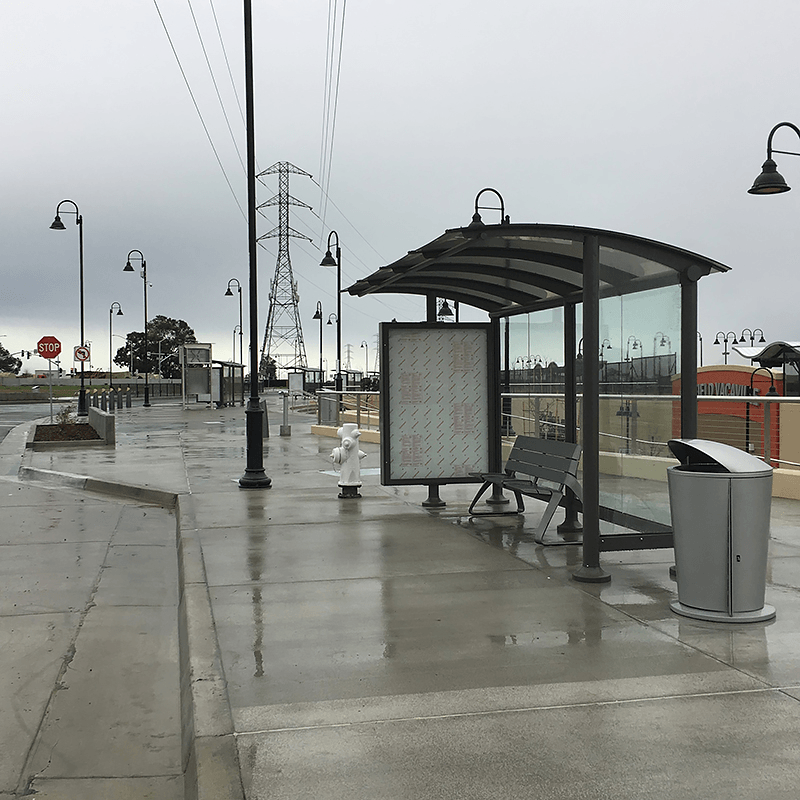 A siga sunset bus shelter is seen from the right side in the rain with a covered bus shelter with bench seating and a double sided advertising display.