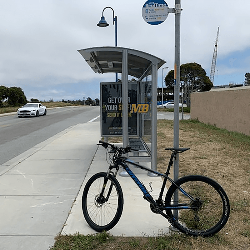 A siga sunset bus shelter with glass siding is seen from the right side, a custom MB logo, and bench seating for three and a bike outside the shelter leaning against the bus stop sign.