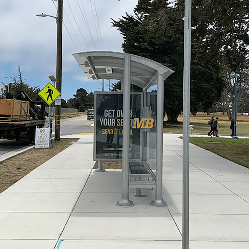 A siga sunset bus shelter with glass siding is seen from the right side, with a custom MB logo, an advertising display on the left. panel, and bench seating for three.