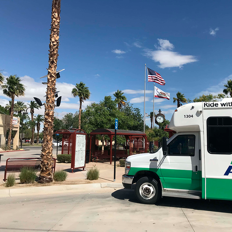 A bus pulls up to a bus stop labeled 3 with bus shelters in the background, benches and a rider information display kiosk.
