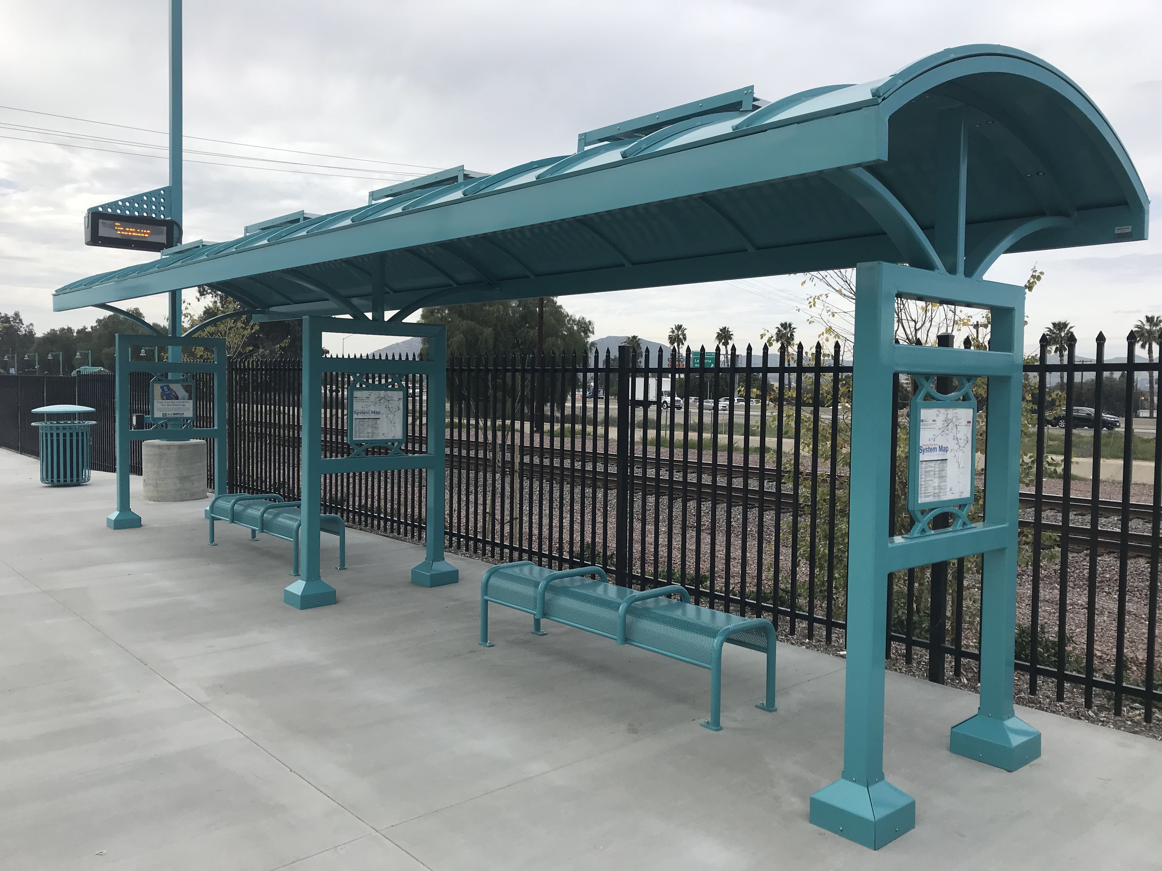 A signa custom bus shelter is seen from the front right with two shelters next to one another with bench seating under each alongside train tracks in the background.