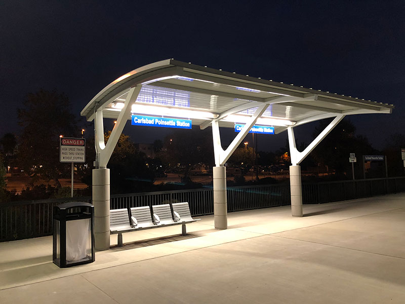 A bus shelter at night seen from the front left with a bench and trash receptacle.