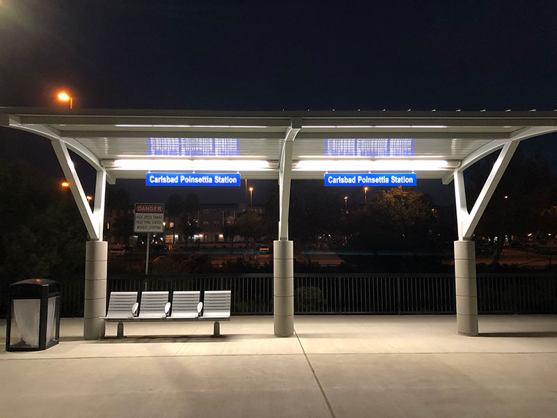 A bus shelter seen from the front with a lit up shelter with bench seating and a trash receptacle on the outside left.