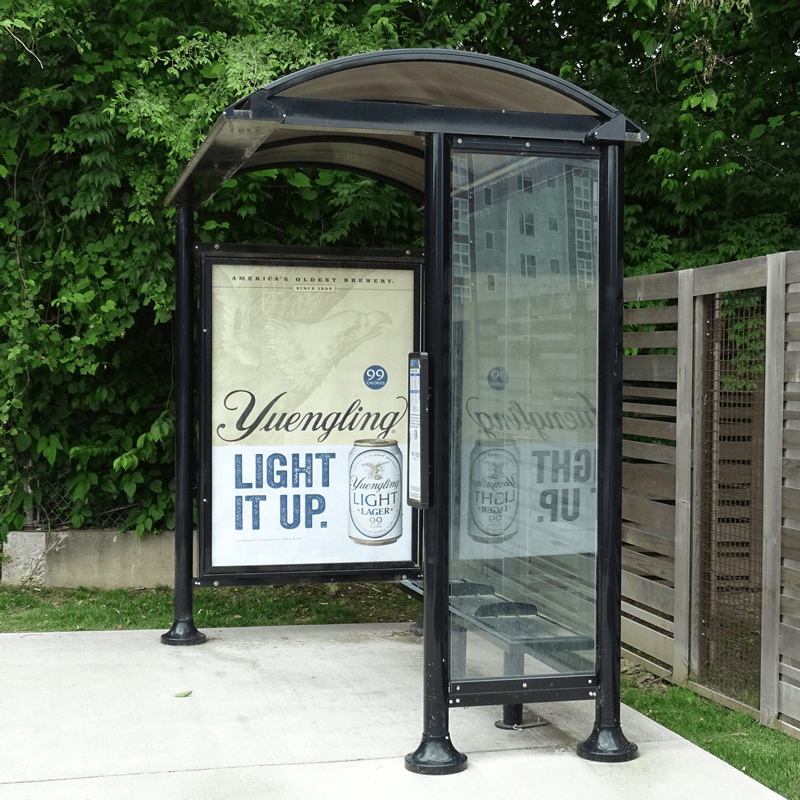 A bus shelter seen from the right side with a left panel advertisement display, bench seating and glass paneling.