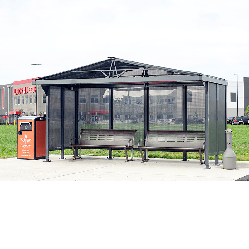 A bus shelter with a pitched roof, two benches for seating inside the shelter, a trash receptacle on the left side and a cigarette receptacle on the right side.