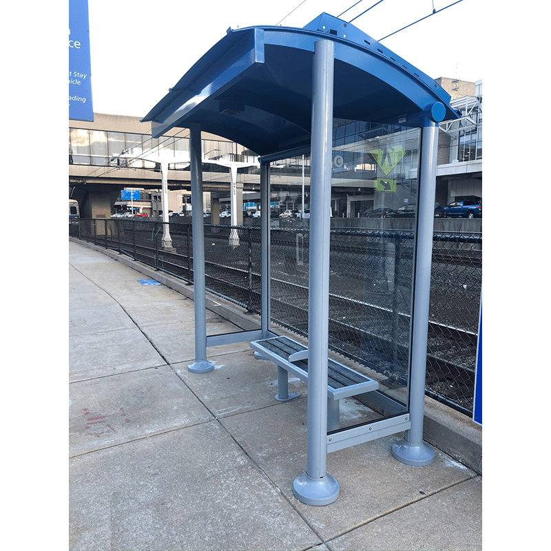 A transit shelter is seen from the front right alongside a set of tracks with a bench for two and glass siding.