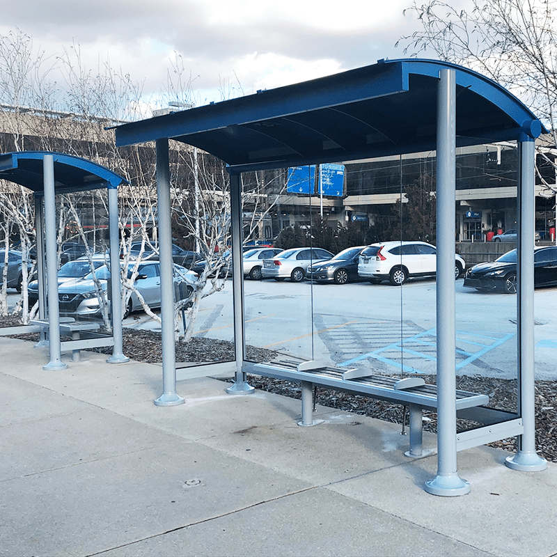 Two bus shelters are seen side by side in front of a parking lot with bench seating and glass side paneling.