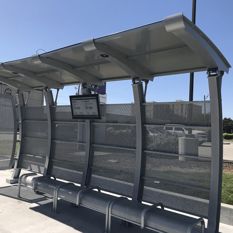A signa crescent bus shelter with bench seating and a digital rider information display on the back showing the time until the next buses arrive.
