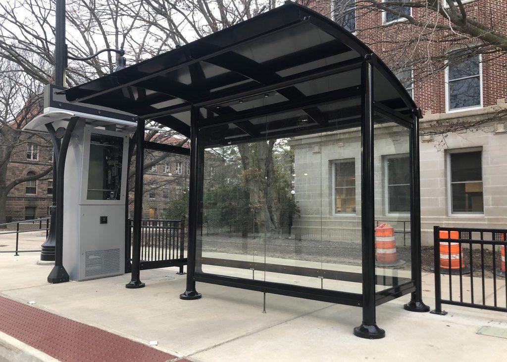 A four sided bus shelter with one entrance with glass walls and a digital display on the right.
