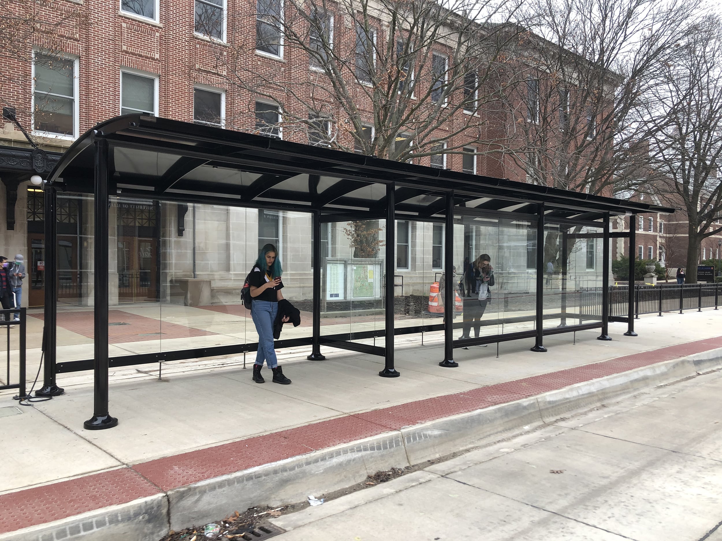 Two riders are seen standing in two bus shelters separated by an enclosed shelter and an open shelter.