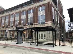 a front stepped back view of a bus shelter with four sides of glass siding, an entrance for passengers and a kiosk for information to the right